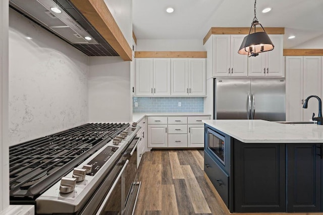 kitchen with wall chimney range hood, white cabinetry, a sink, and built in appliances