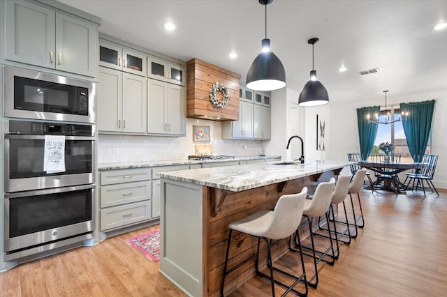 kitchen featuring double oven, black microwave, an island with sink, sink, and light stone countertops