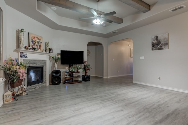 living room with a stone fireplace, a tray ceiling, ceiling fan, and light hardwood / wood-style flooring