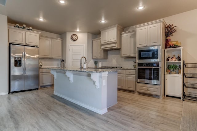 kitchen with light hardwood / wood-style flooring, a kitchen island with sink, stainless steel appliances, light stone countertops, and a kitchen bar