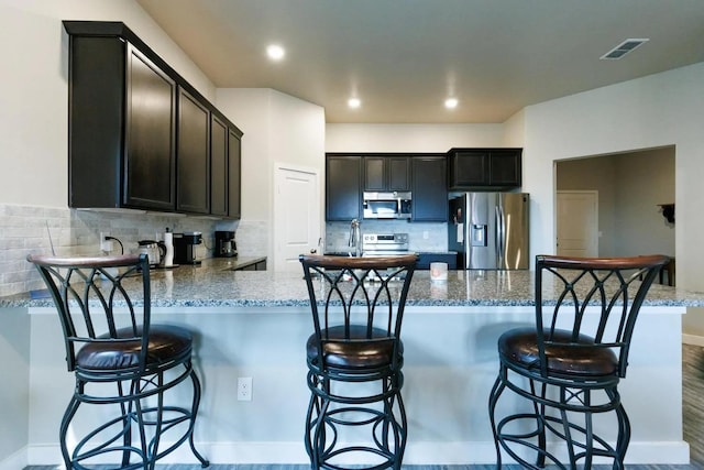 kitchen featuring dark brown cabinetry, a breakfast bar area, light stone counters, stainless steel appliances, and decorative backsplash