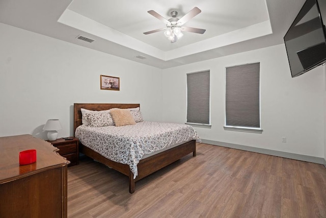 bedroom featuring a ceiling fan, wood finished floors, visible vents, baseboards, and a tray ceiling