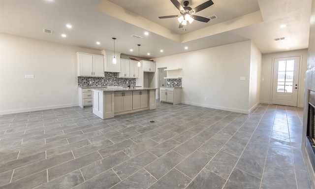 kitchen featuring pendant lighting, tasteful backsplash, an island with sink, white cabinets, and a raised ceiling