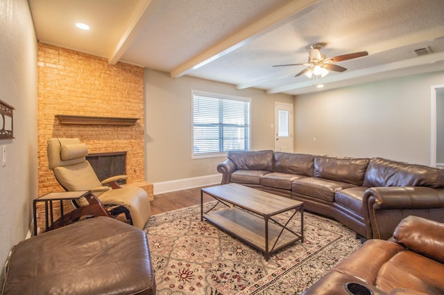 living room with ceiling fan, beam ceiling, wood-type flooring, a textured ceiling, and a brick fireplace