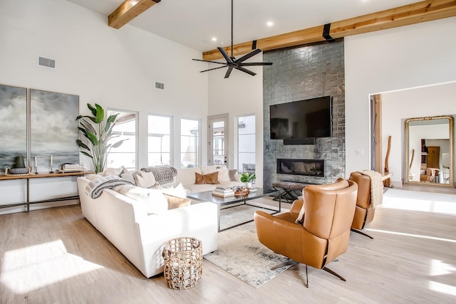 living room with light wood-type flooring, ceiling fan, a large fireplace, beamed ceiling, and a towering ceiling