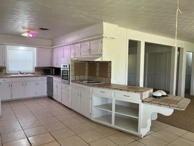 kitchen featuring light tile patterned floors, appliances with stainless steel finishes, white cabinetry, tile counters, and kitchen peninsula