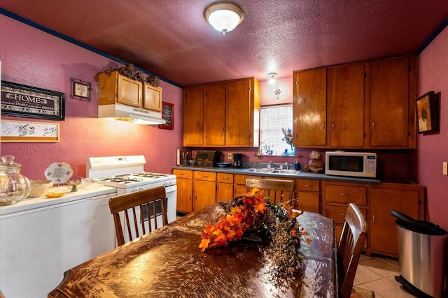 kitchen with dark countertops, under cabinet range hood, brown cabinets, white appliances, and a sink