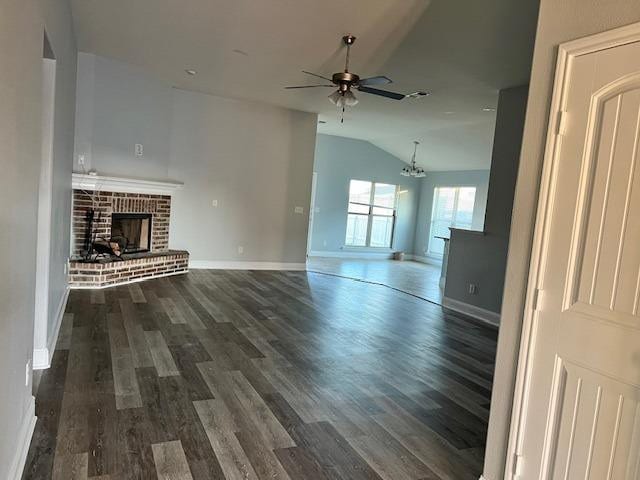 unfurnished living room with dark wood-type flooring, a brick fireplace, vaulted ceiling, baseboards, and ceiling fan with notable chandelier