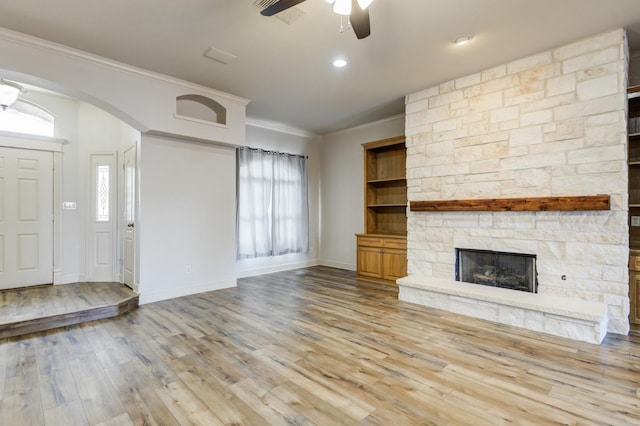 unfurnished living room featuring a stone fireplace, crown molding, light hardwood / wood-style flooring, built in features, and ceiling fan