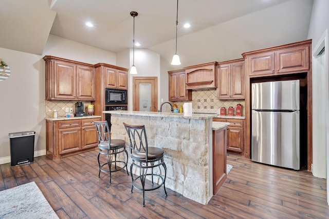 kitchen with premium range hood, hanging light fixtures, black appliances, light stone countertops, and a center island with sink