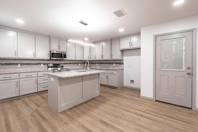 kitchen featuring appliances with stainless steel finishes, pendant lighting, an island with sink, light stone counters, and light wood-type flooring