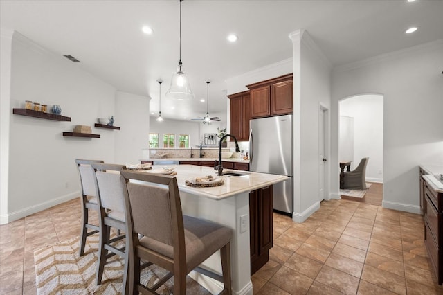 kitchen with a breakfast bar area, hanging light fixtures, stainless steel refrigerator, ornamental molding, and an island with sink