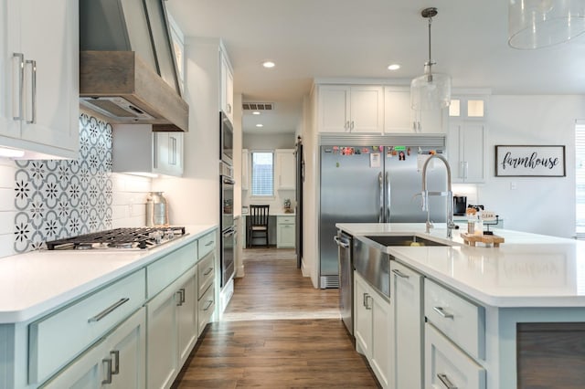 kitchen featuring white cabinets, hanging light fixtures, built in appliances, custom range hood, and a center island with sink