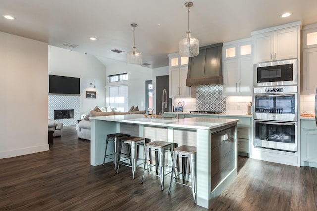 kitchen with custom range hood, white cabinets, a center island with sink, built in microwave, and decorative light fixtures