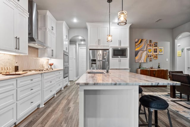 kitchen featuring hanging light fixtures, wall chimney range hood, an island with sink, stainless steel appliances, and white cabinets