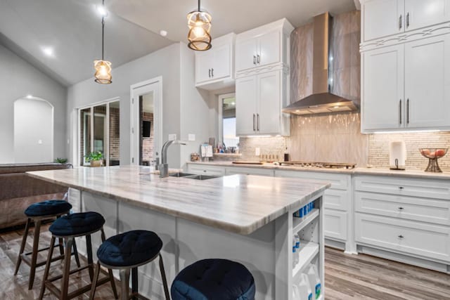 kitchen with white cabinetry, a kitchen island with sink, wall chimney range hood, and decorative light fixtures