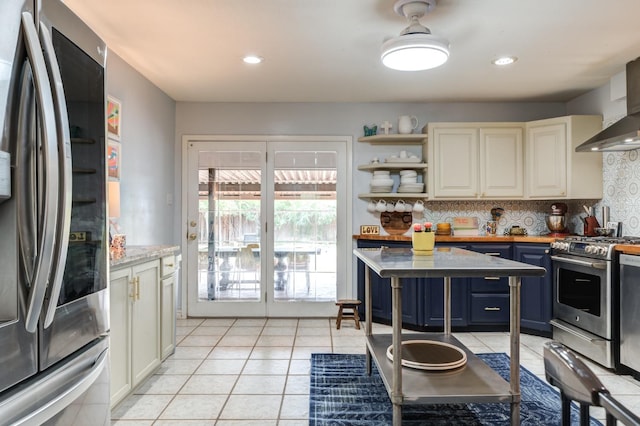 kitchen featuring light tile patterned floors, wall chimney range hood, blue cabinetry, appliances with stainless steel finishes, and decorative backsplash