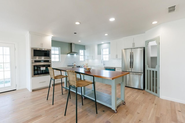 kitchen with appliances with stainless steel finishes, tasteful backsplash, white cabinets, light hardwood / wood-style floors, and wall chimney range hood