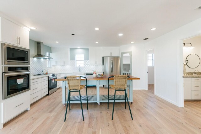 kitchen featuring appliances with stainless steel finishes, tasteful backsplash, white cabinets, wall chimney exhaust hood, and light hardwood / wood-style flooring