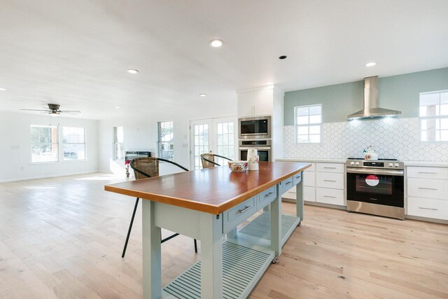 kitchen with appliances with stainless steel finishes, tasteful backsplash, white cabinets, wall chimney range hood, and light wood-type flooring