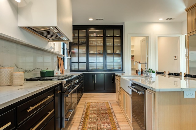 kitchen featuring sink, stainless steel appliances, a center island with sink, exhaust hood, and light wood-type flooring