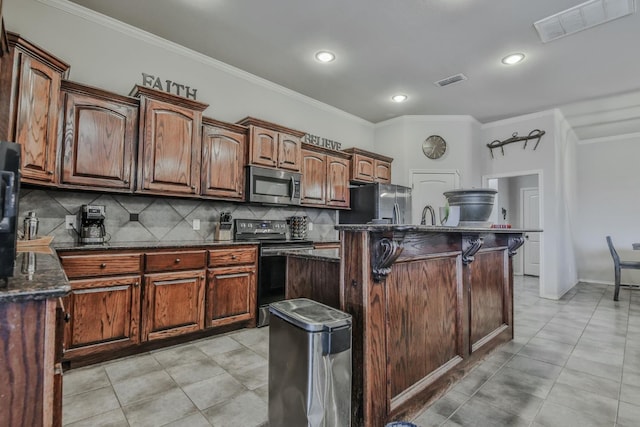 kitchen featuring crown molding, dark stone countertops, an island with sink, stainless steel appliances, and decorative backsplash