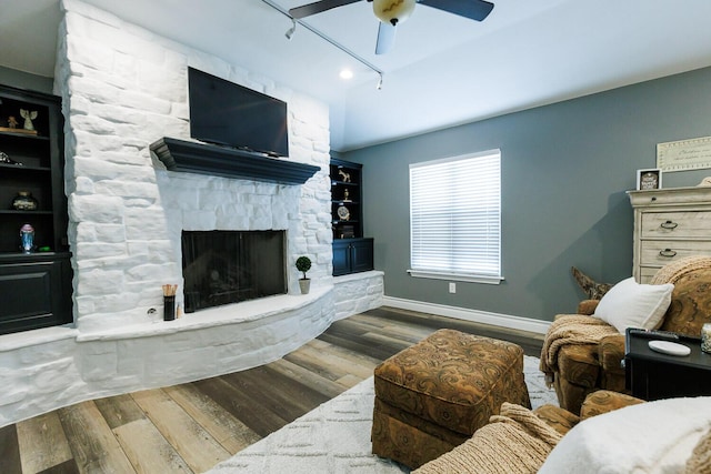 living room with track lighting, a ceiling fan, wood finished floors, a stone fireplace, and baseboards