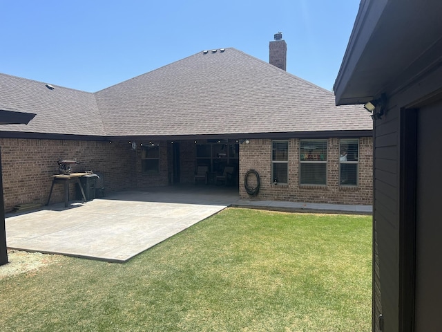 rear view of house featuring a patio, a shingled roof, brick siding, a lawn, and a chimney