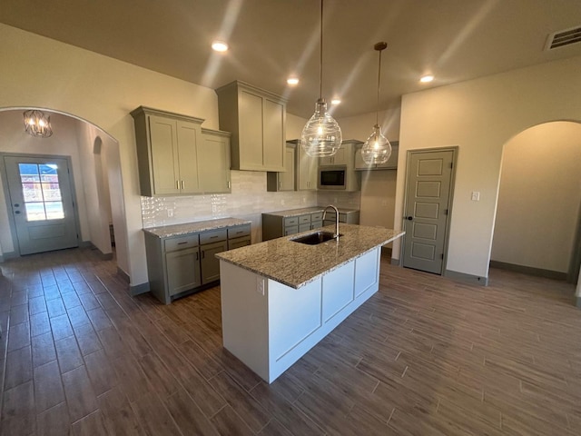 kitchen featuring sink, light stone counters, decorative light fixtures, stainless steel microwave, and an island with sink