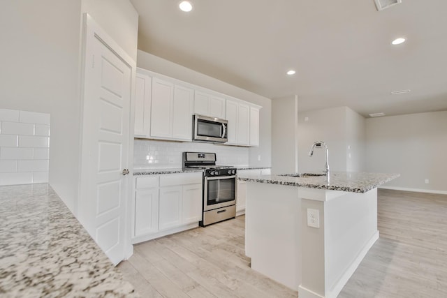 kitchen featuring light stone counters, light wood-type flooring, appliances with stainless steel finishes, an island with sink, and white cabinets