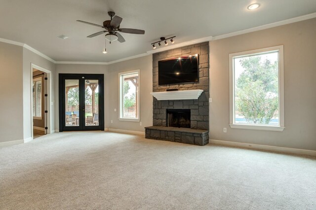 unfurnished living room featuring a stone fireplace, light colored carpet, ceiling fan, crown molding, and french doors