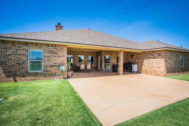 back of house featuring roof with shingles, a chimney, a patio area, brick siding, and a lawn