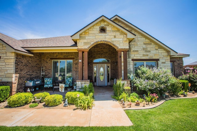 view of exterior entry featuring roof with shingles, stone siding, a porch, brick siding, and a lawn