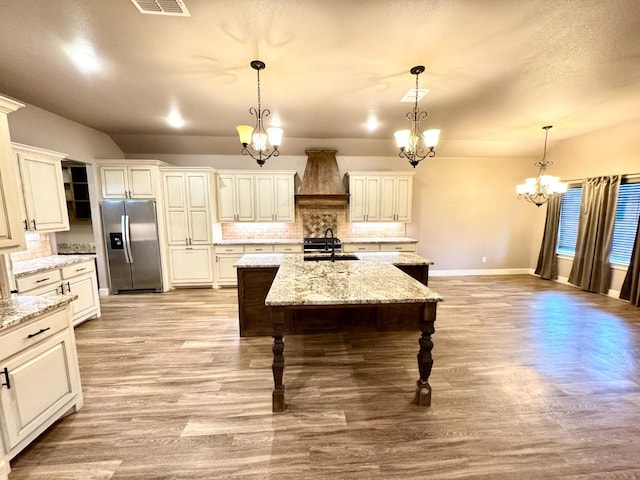 kitchen with pendant lighting, sink, stainless steel fridge, custom range hood, and a center island with sink