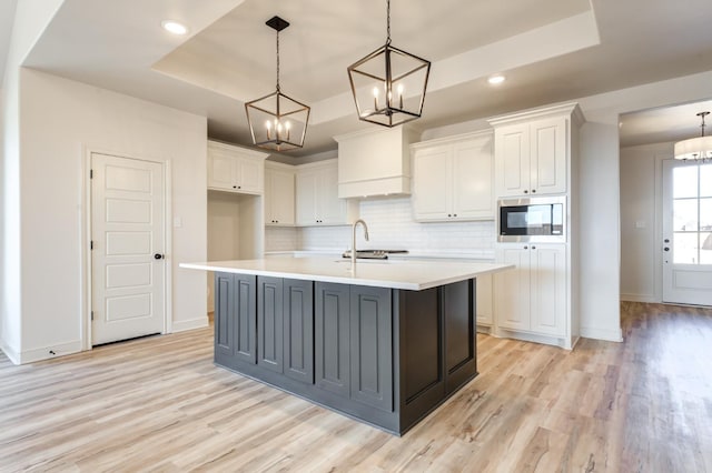 kitchen with pendant lighting, stainless steel microwave, a tray ceiling, an island with sink, and white cabinets