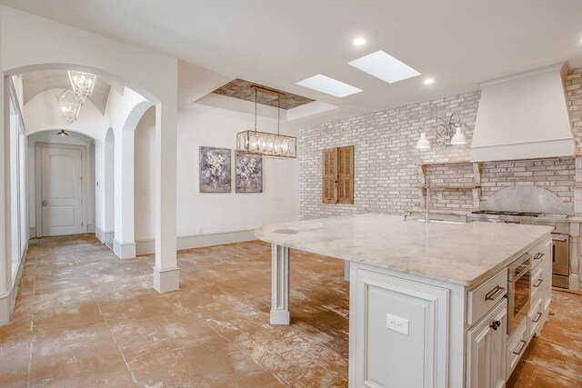 kitchen featuring decorative light fixtures, light stone countertops, custom range hood, a center island with sink, and stainless steel range oven