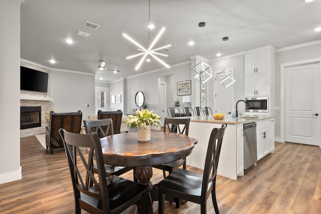 dining room with sink, crown molding, ceiling fan with notable chandelier, a stone fireplace, and light wood-type flooring