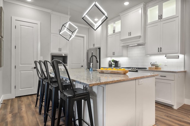 kitchen with appliances with stainless steel finishes, white cabinetry, an island with sink, ornamental molding, and light stone counters