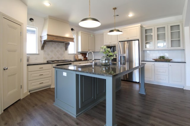 kitchen with custom exhaust hood, hanging light fixtures, dark hardwood / wood-style flooring, stainless steel appliances, and a kitchen island with sink