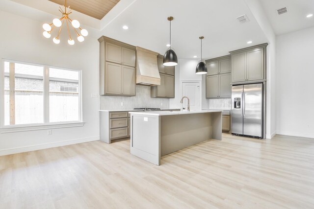 kitchen with tasteful backsplash, decorative light fixtures, appliances with stainless steel finishes, an island with sink, and custom range hood