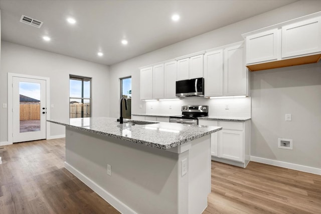 kitchen featuring white cabinets, backsplash, stainless steel appliances, light stone countertops, and a center island with sink