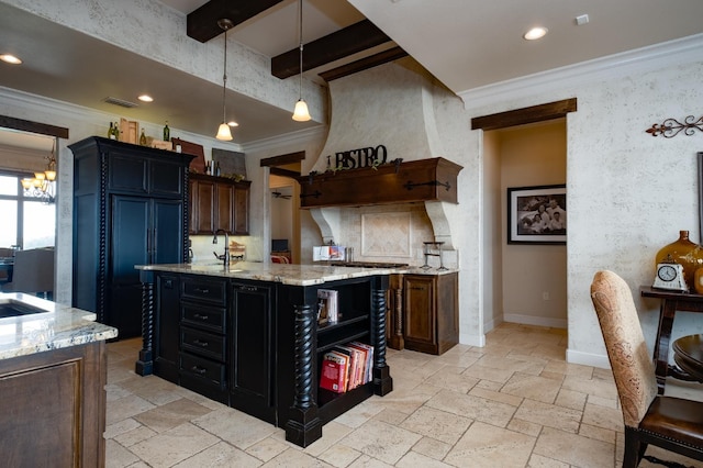 kitchen with light stone counters, hanging light fixtures, ornamental molding, and a center island with sink