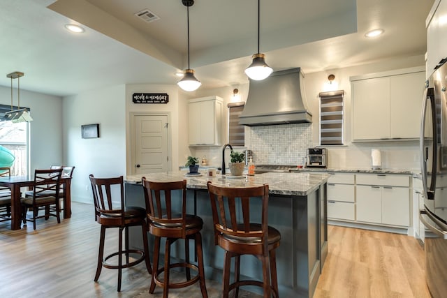 kitchen featuring an island with sink, custom range hood, white cabinets, pendant lighting, and backsplash