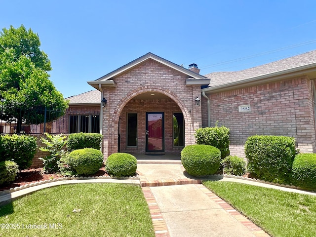 9802 Toledo Ave, Lubbock, TX 79424 - property entrance featuring brick siding, a chimney, a yard, and a shingled roof