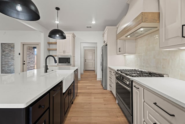 kitchen with appliances with stainless steel finishes, white cabinetry, custom range hood, an island with sink, and decorative light fixtures