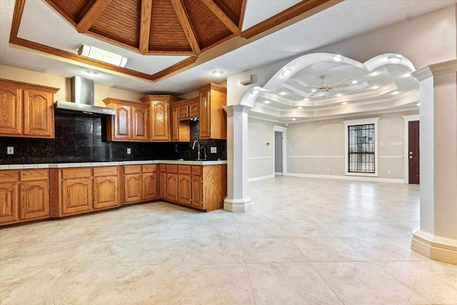 kitchen featuring wall chimney range hood, a tray ceiling, sink, and ornate columns