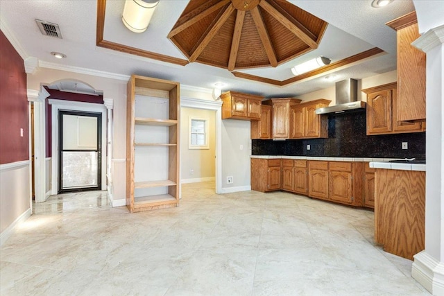 kitchen featuring wall chimney range hood, crown molding, decorative columns, a tray ceiling, and tile counters