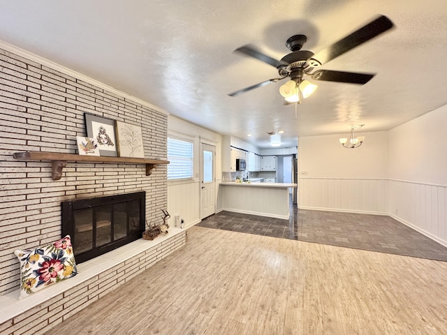 unfurnished living room with a textured ceiling, a brick fireplace, wainscoting, ceiling fan with notable chandelier, and dark wood-type flooring
