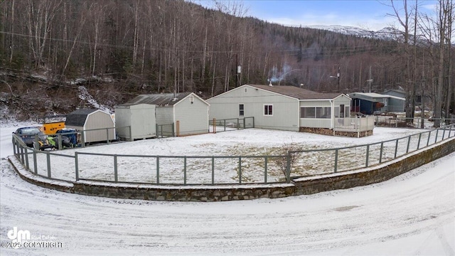 snow covered house with a storage shed, an outbuilding, fence, and a forest view