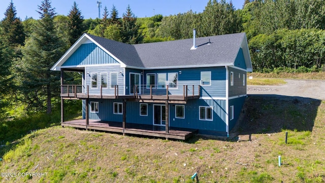 back of house with a shingled roof, a deck, a view of trees, and a yard